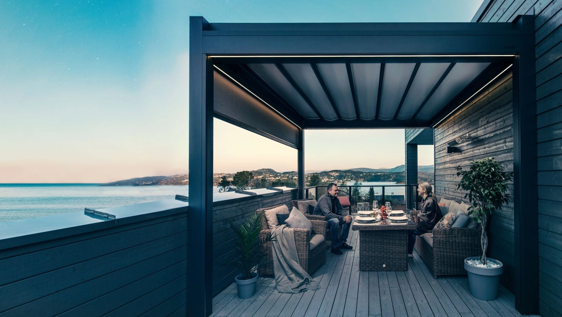 Ein Mann und eine Frau sitzen auf einem Balkon am Meer, überdacht von dem Textildach Brustor B300.