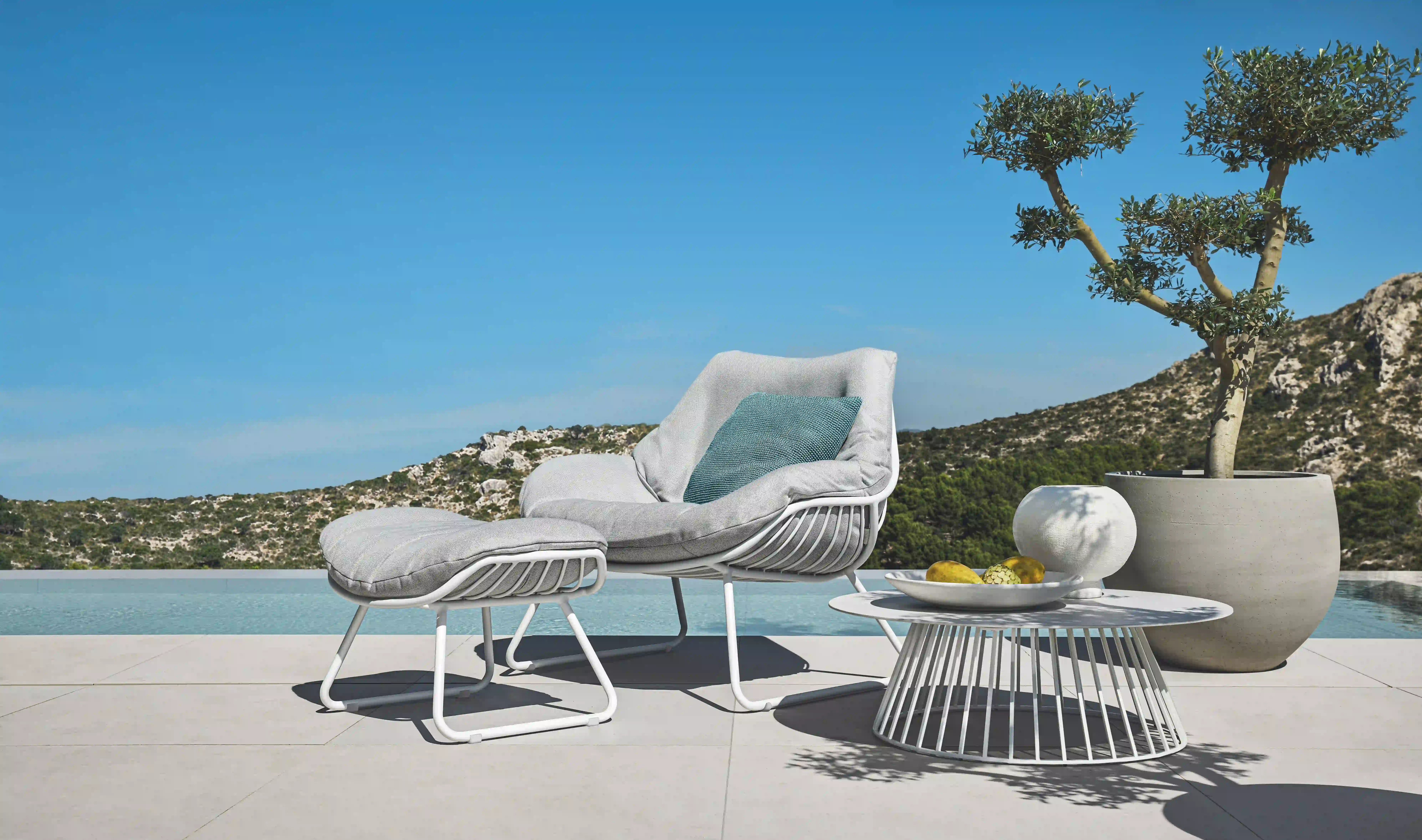 Aus dem Sortiment der Terrassenmöbel von Solpuri werden hier ein CHILL Lounge Sessel XL mit Hocker und Beistelltisch GRID vor einem Pool mit weiter Aussicht kombiniert.