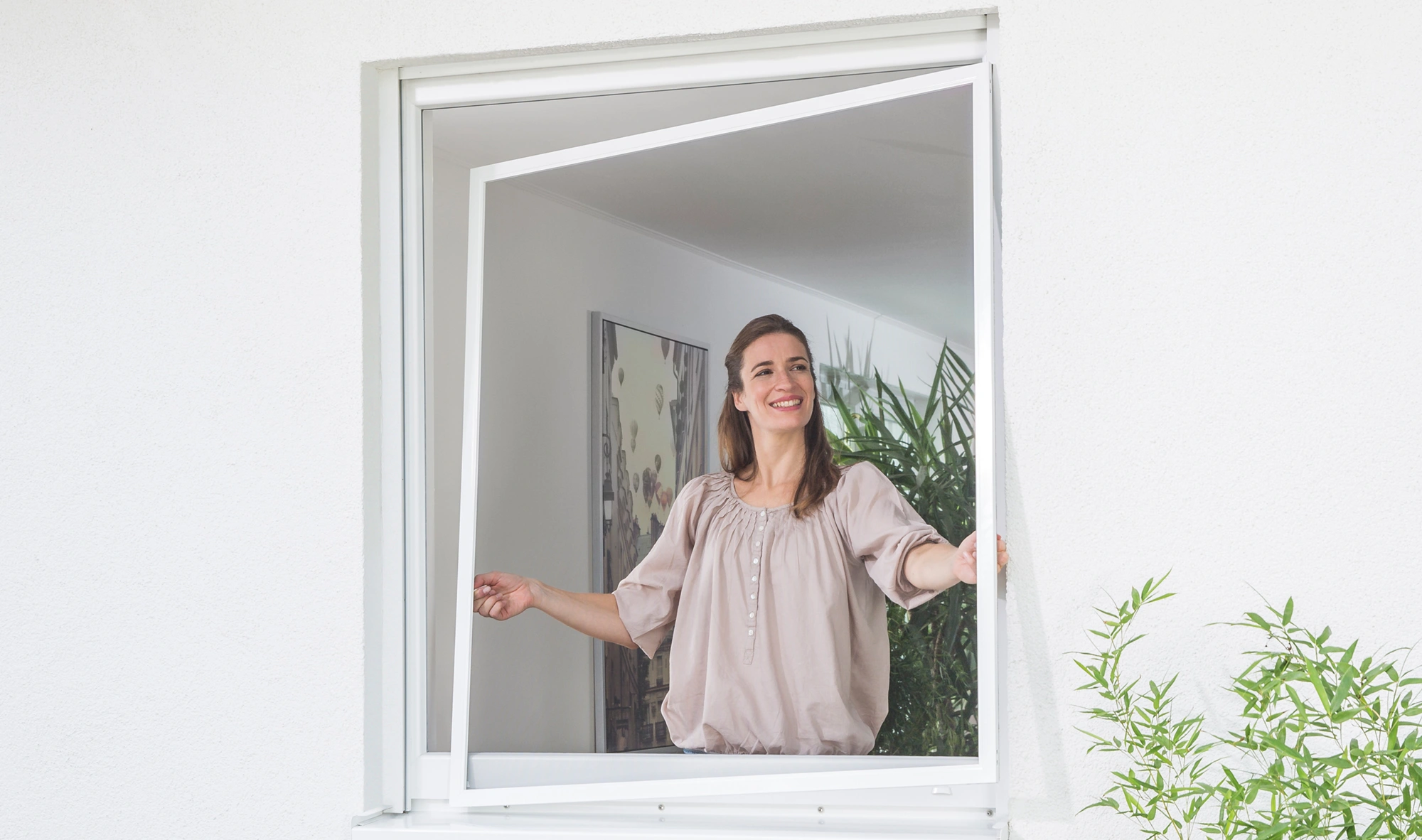 Eine Frau mit transparentem Insektenschutzgitter mit weißem Rahmen, den sie in ein Fenster gleicher Farbe einsetzt.