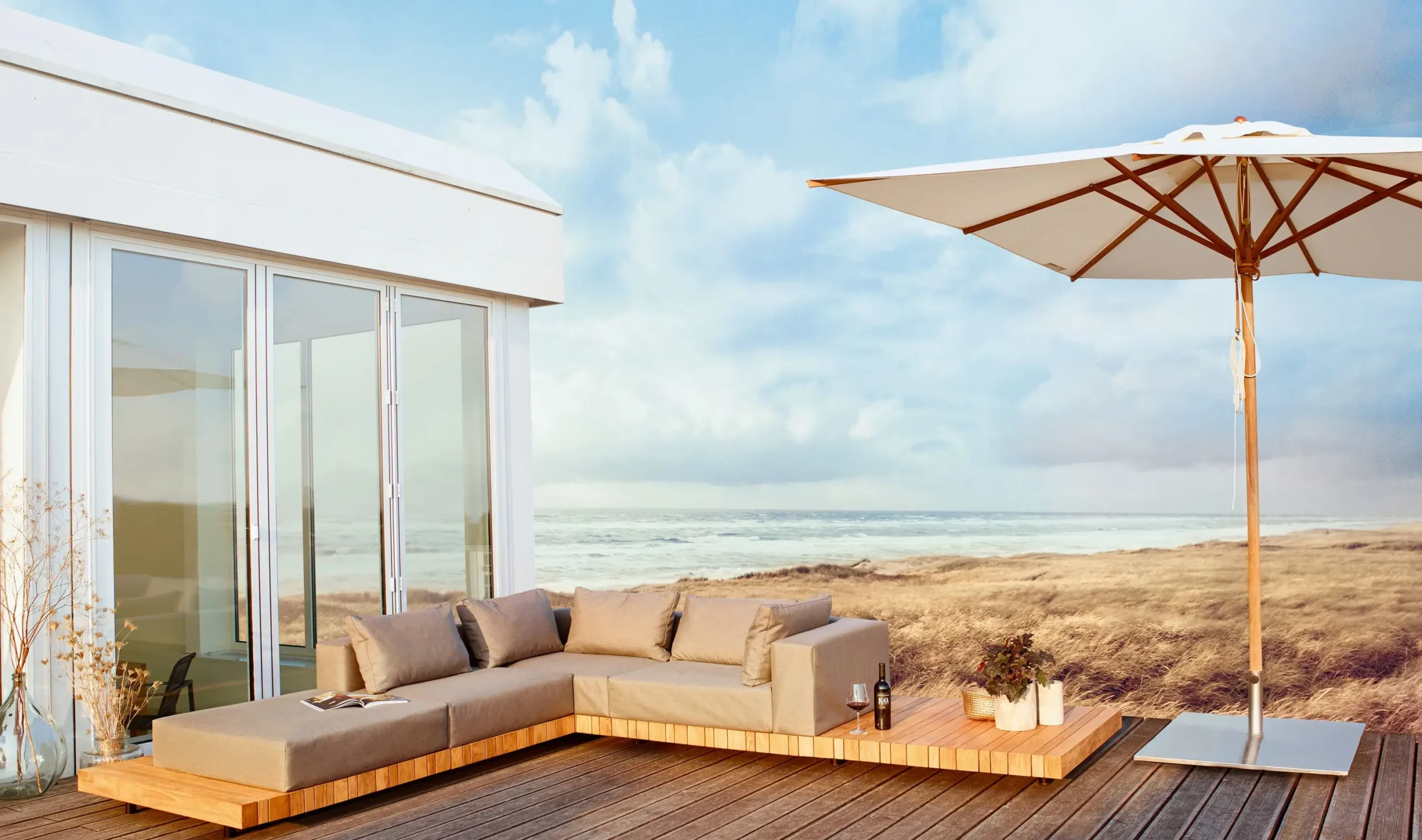 Eine PLATEAU Lounge der Solpuri Terrassenmöbel auf dem Holzdeck eines Strandhauses vor Dünen und Meer.