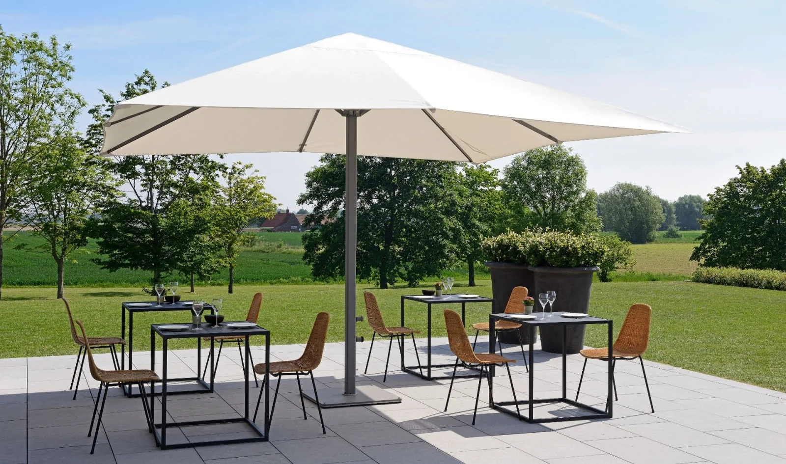 Ein weißer Prostor P8 Sonnenschirm beschattet vier Zweiertische auf einer Terrasse vor einer Wiese mit Bäumen.