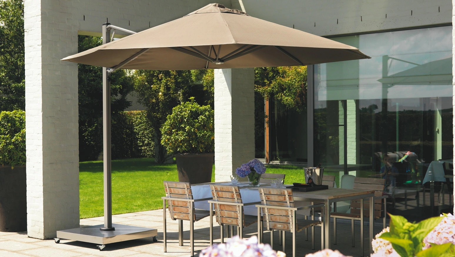 Ein Prostor P7 Sonnenschirm in bräunlicher Farbe über einem Esstisch auf der Terrasse eines modernen Hauses mit Garten.