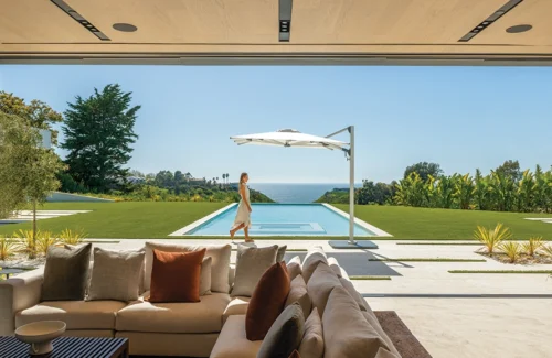 Ein Shademaker Solaris beschattet einen Pool in der Wiese eines Hauses mit Meerblick. Auf der Terrasse davor läuft eine Frau.