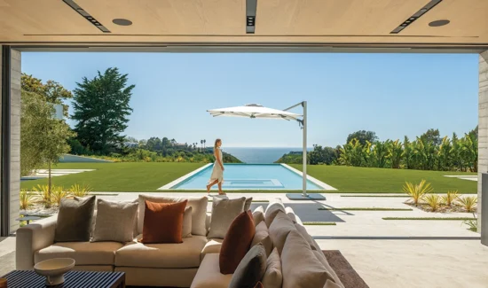 Ein Shademaker Solaris beschattet einen Pool in der Wiese eines Hauses mit Meerblick. Auf der Terrasse davor läuft eine Frau.