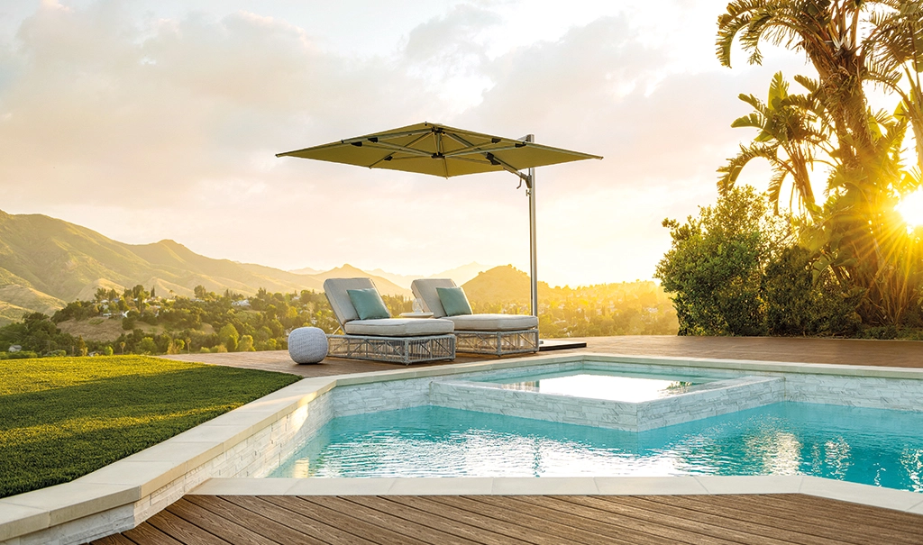 Ein eckiger Shademaker Solaris über zwei Liegen an einem Pool mit Bergen im Hintergrund.