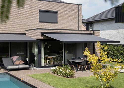 Eine Hausfassade in beigem Ziegel. zwei Markisen in dunkelgrau. Ein Teil Rasen und Terrasse.
