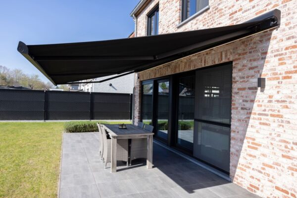 Eine ausgefahrene Markise mit schwarzem Stoff und schwarzem Gestell ist an einer Hauswand rechts im Bild befestigt und beschattet einen Essplatz auf einer Terrasse darunter.
