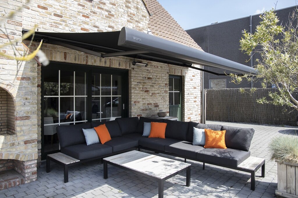 eine Markise mit schwarzem Gestell und schwarzem Stoff ist über einer Sitzgruppe mit antrazitfarbenem Stoff und grauen und orangenen Kissen ausgefahren und beschattet diese