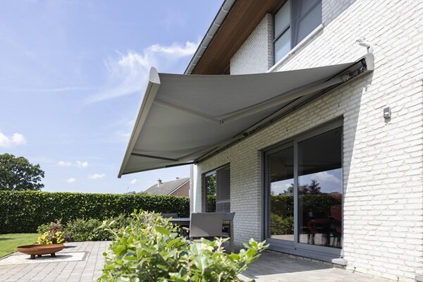 Eine hellgraue Markise ist über einem Esstisch auf der Terrasse und den dahinter liegenden großen Terrassenfenstern ausgefahren.